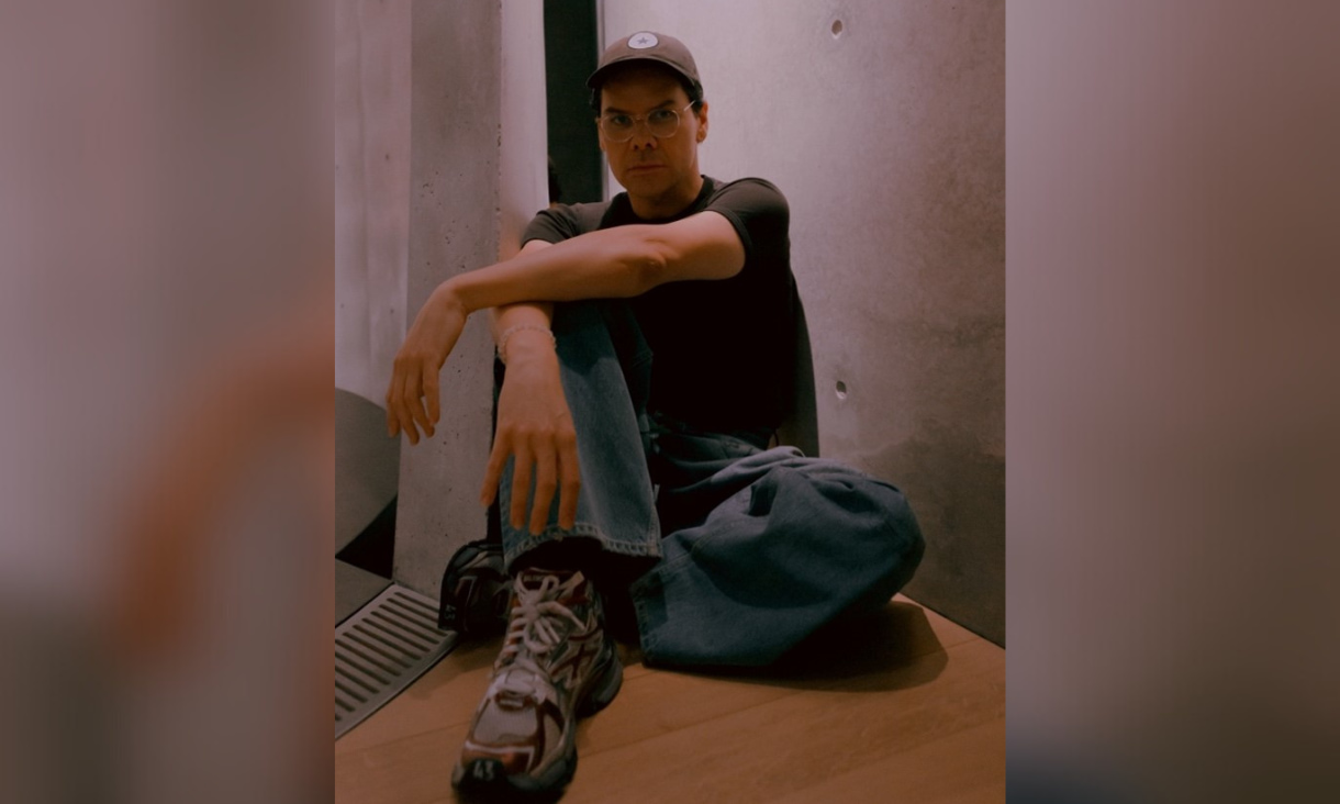 Christian Thompson sitting on the ground in front of a blank wall. He's wearing a hat, a black shirt and jeans.